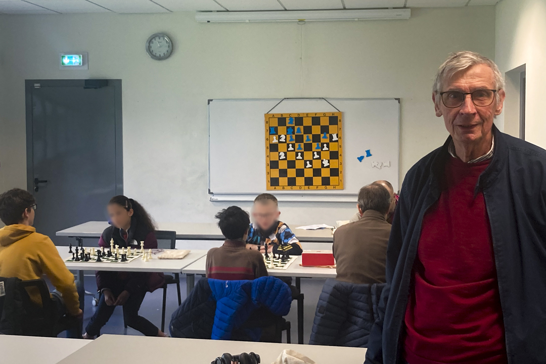 Claude, président de l'Échiquier Bisontin lors d'un cours d'échecs à la maison de quartier Nelson Mandela. Il pause face caméra pour un portrait dans une salle de classe, derrière lui des adultes et enfants jouent aux échecs.