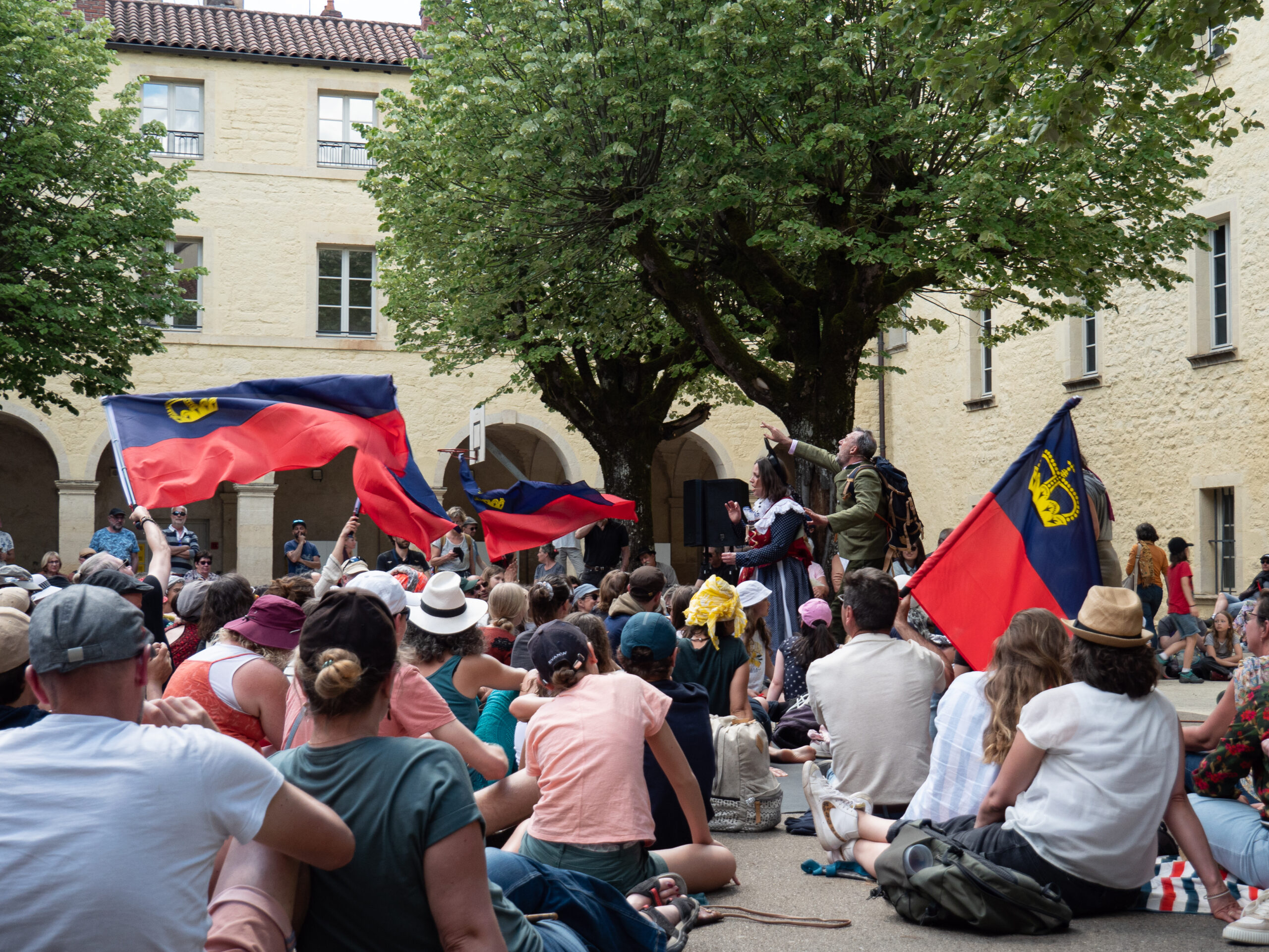 Foule assise en train d'écouter une représentation théâtrale dans la rue, on aperçoit plusieurs drapeaux du Liechtenstein 