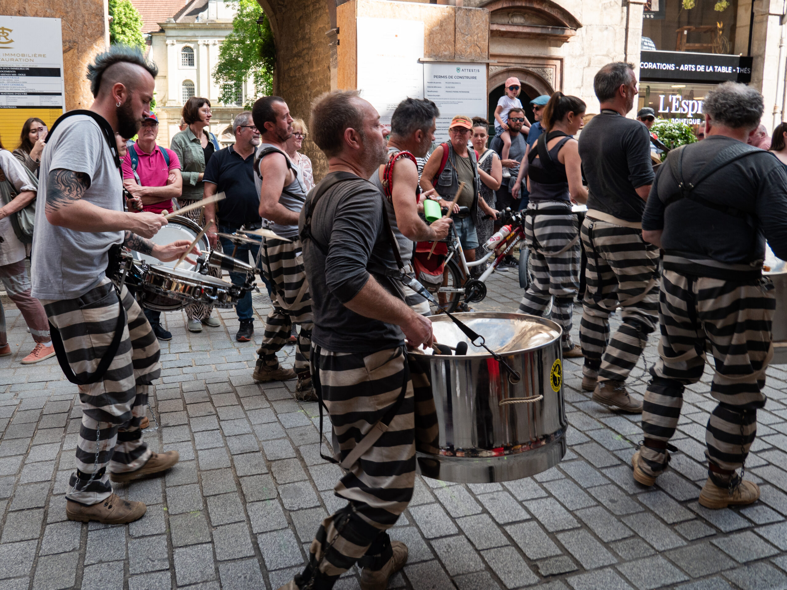 Fanfare de rue composée de diverses percussions, les musiciens sont habillés de bas de pantalon de bagnard, gris à rayures horizontales noires