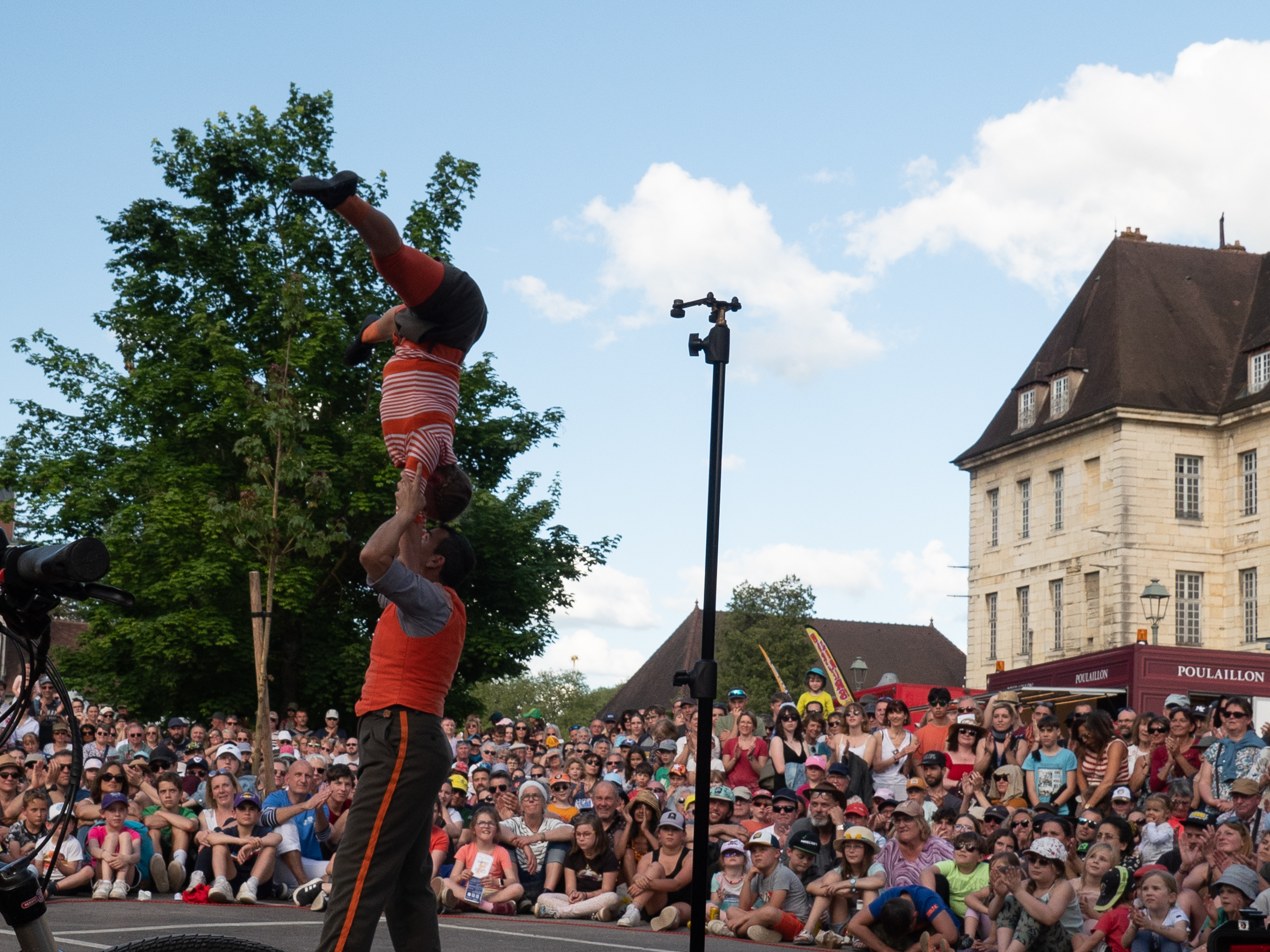 Deux acrobates vêtus de rouge et gris devant une forte foule, la femme est en train de monter sur la tête de l'homme, tête contre tête, en équilibre