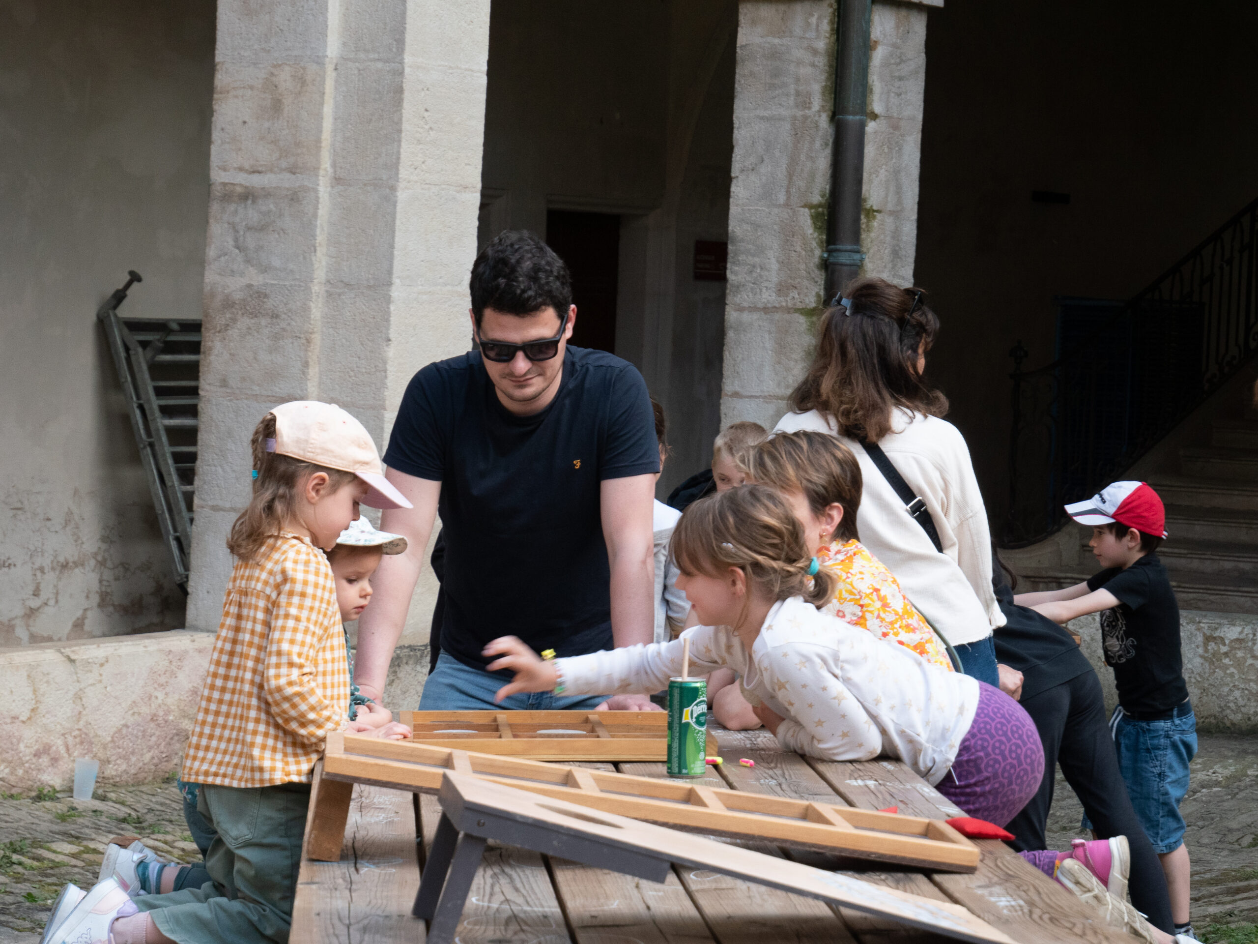 Quatre enfants jouent à un jeu en bois sur une table de pique-nique, surveillés par un homme adulte