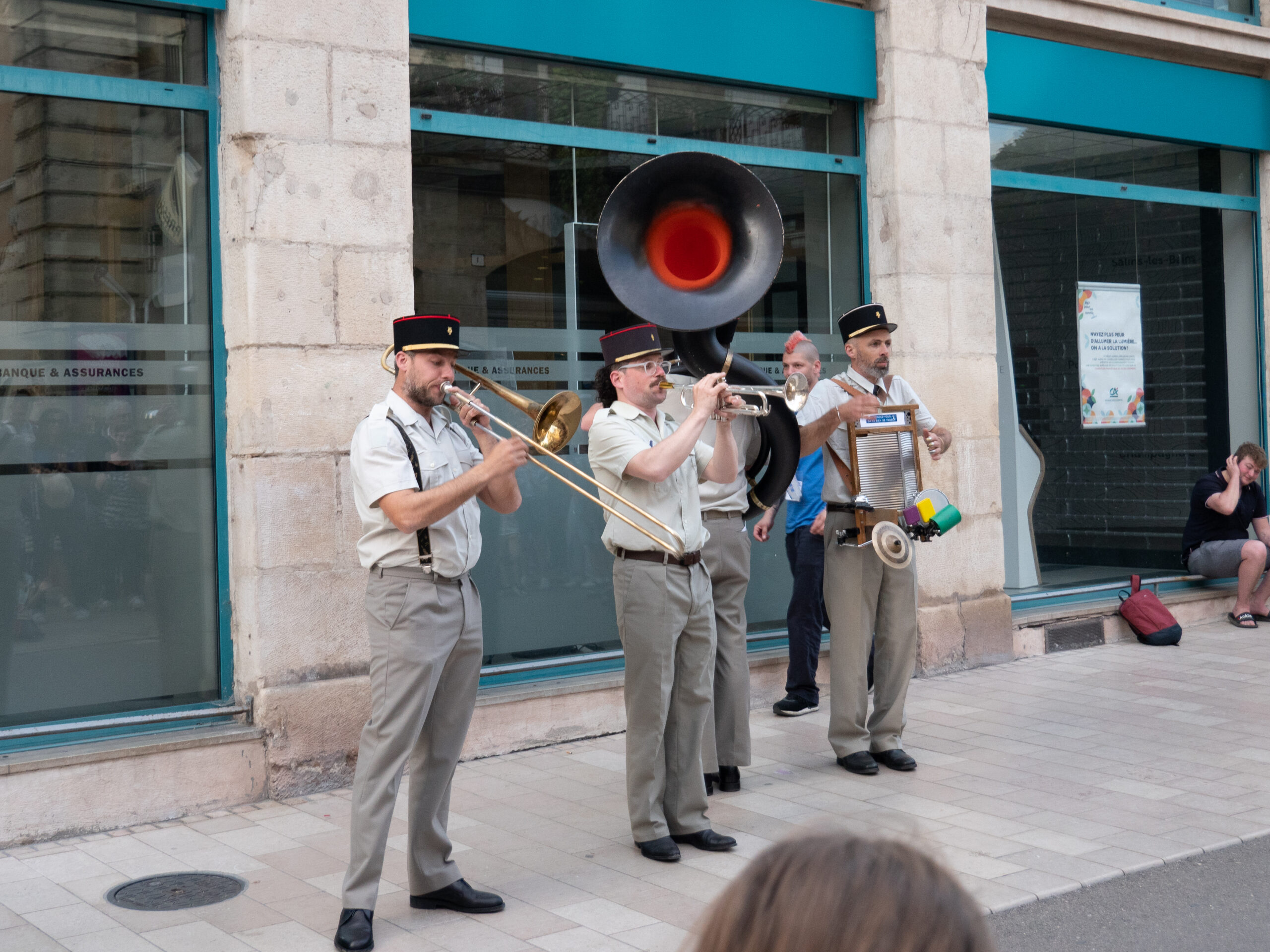quatuor de musiciens habillés en flics, costumes gris et képis, avec un tuba, une trompette, un trombone et un bruiteur / percussionniste, ils sont sur le trottoir