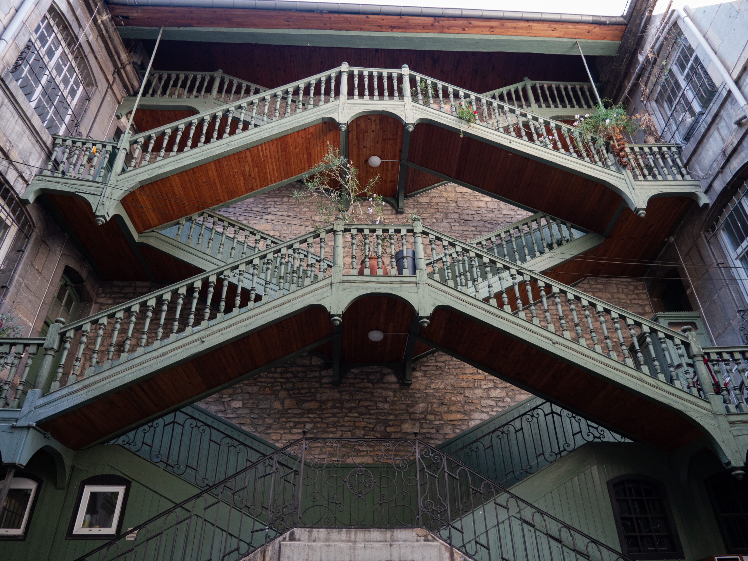 Vue en contre plongée d'un double escalier en bois peint en vert sombre, devant une façade en vieilles pierres, desservant les deux côtés de l'immeuble