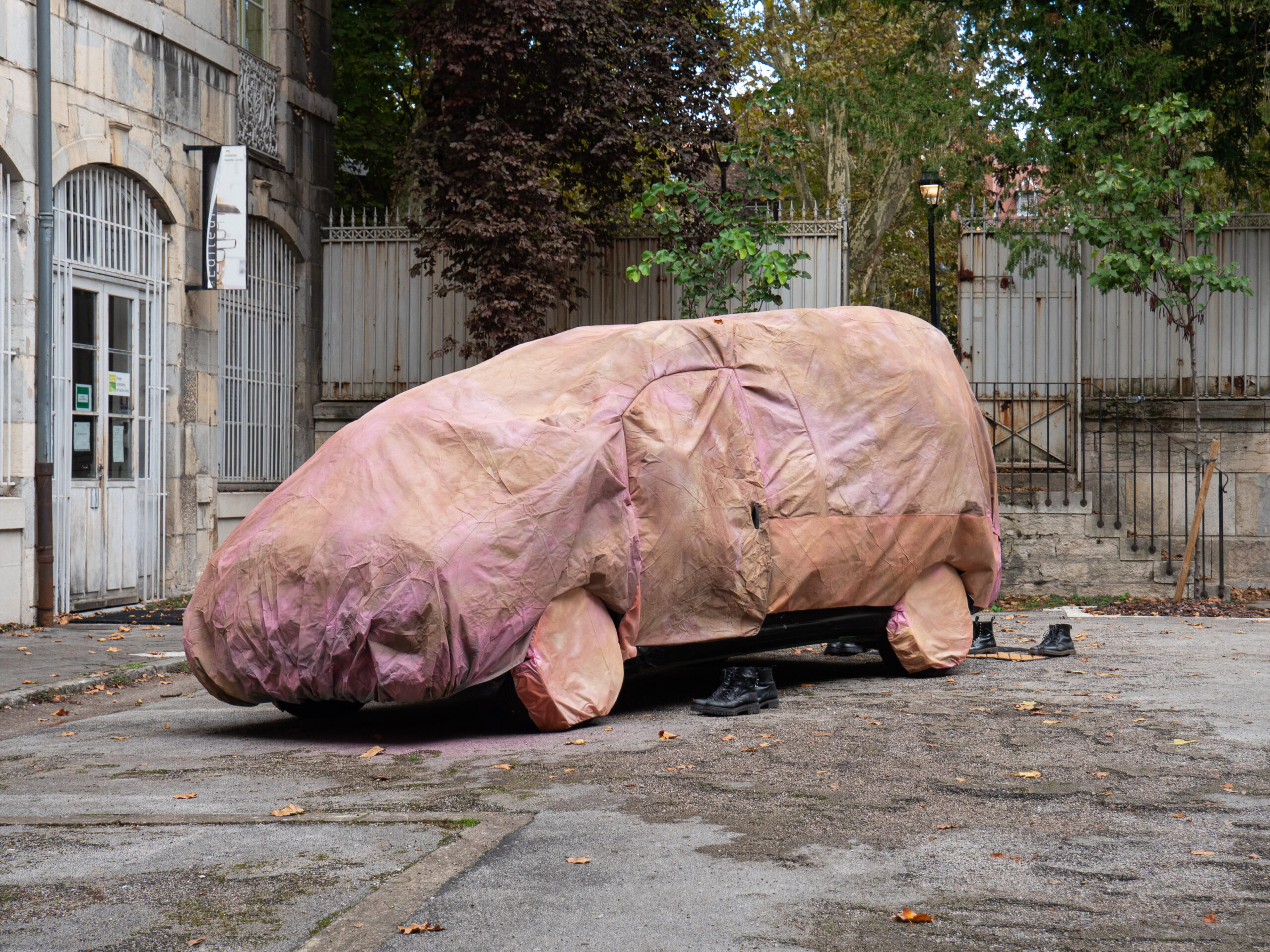 Voiture entièrement entourée et recouverte d'une bâche de draps colorés, autour de laquelle ont été disposées quelques paires de chaussures doc marteens noires