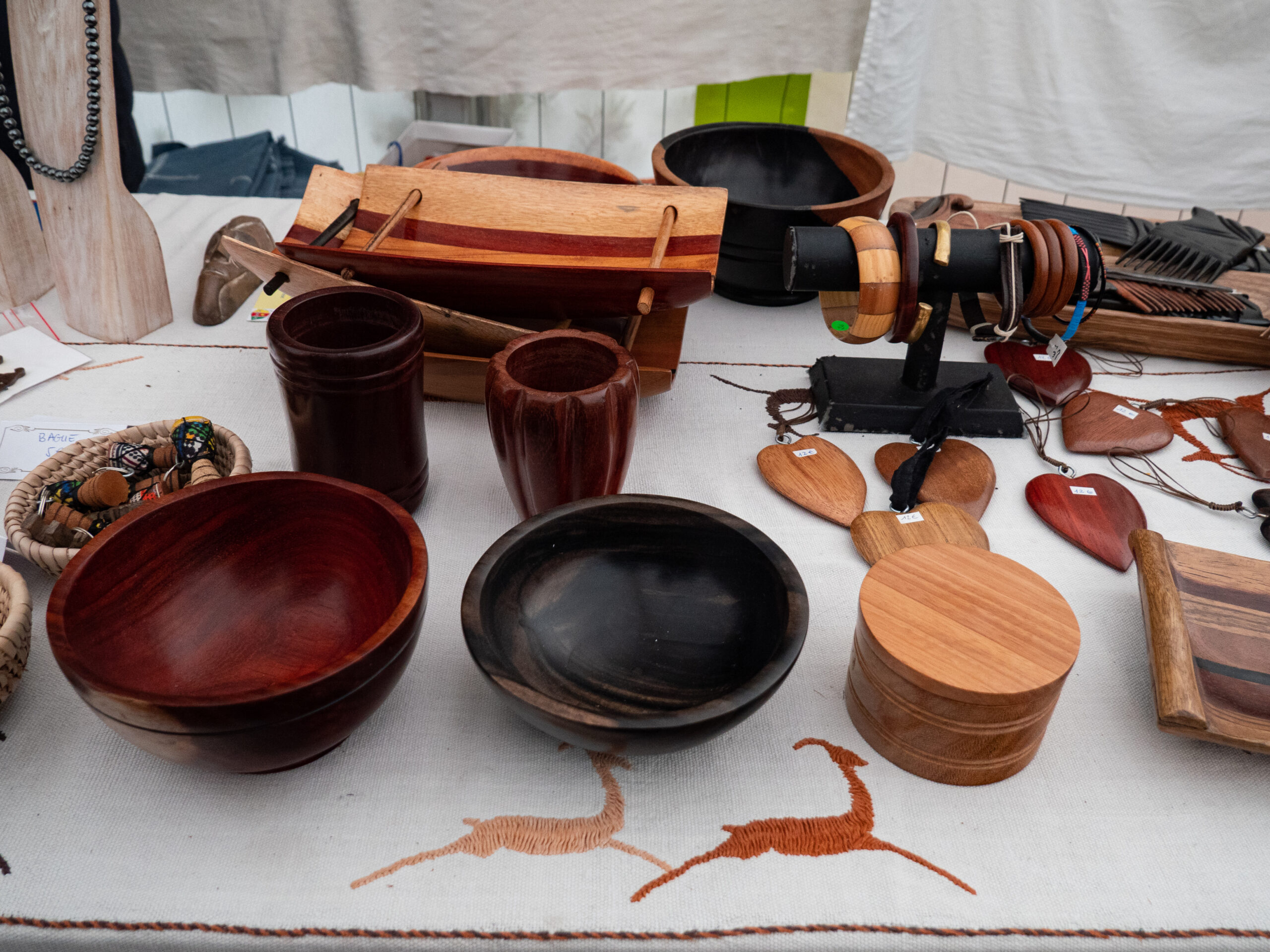 Diverses créations en bois : bols, verres, boite de rangement, pendentifs en cœur, peignes, broches, disposés sur un linge blanc décoré d'antilopes cousues