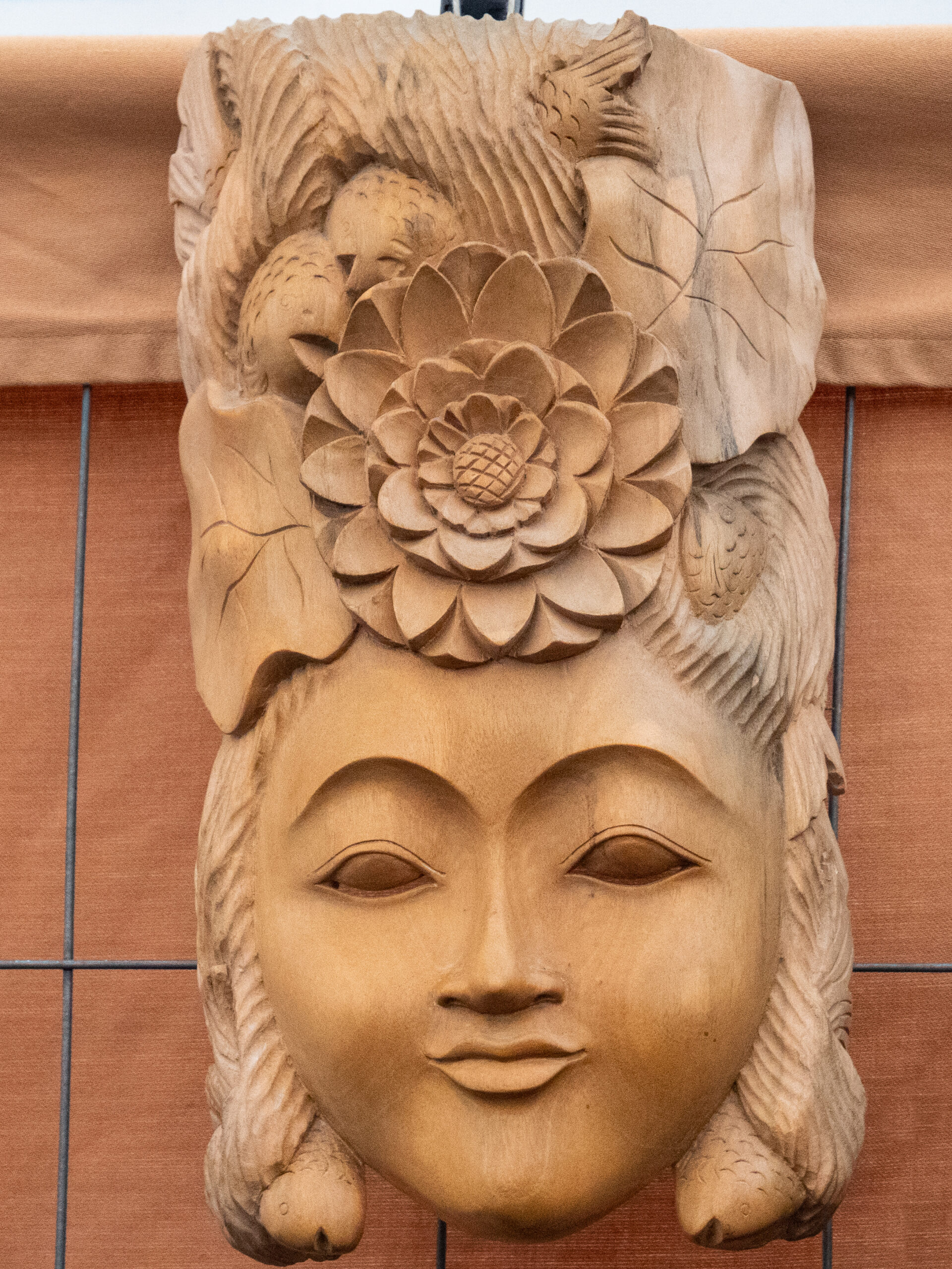 tête de femme souriante sculptée dans du bois, coiffée avec une grosse fleur centrale et divers éléments floraux qui montent