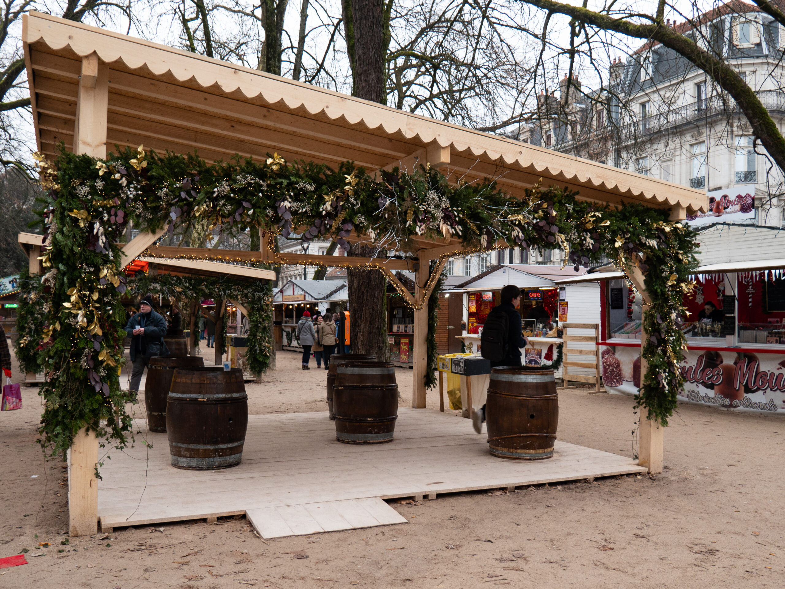 le coin mange-debout du marché de Noël de Besançon, avec de gros tonneaux en guise de table, installés sous un genre de barnum en bois, décoré de branches de sapin et de guirlandes, pris de jour