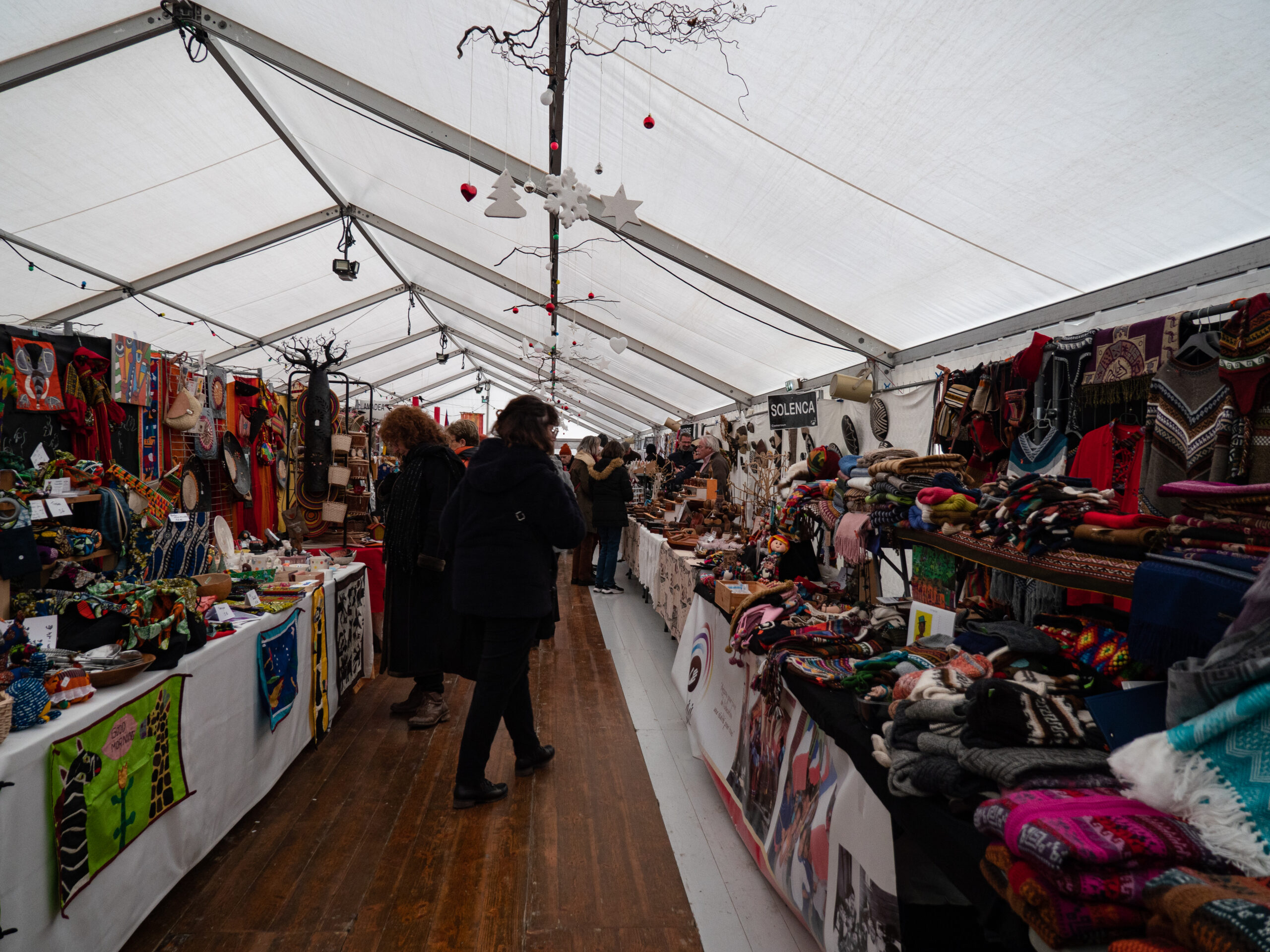 Une allée sous le long chapiteau du marché de Noël solidaire, pleine de stands de tissus, objets artisanaux en bois ou en cuir, devant lesquels quelques personnes sont arrêtées