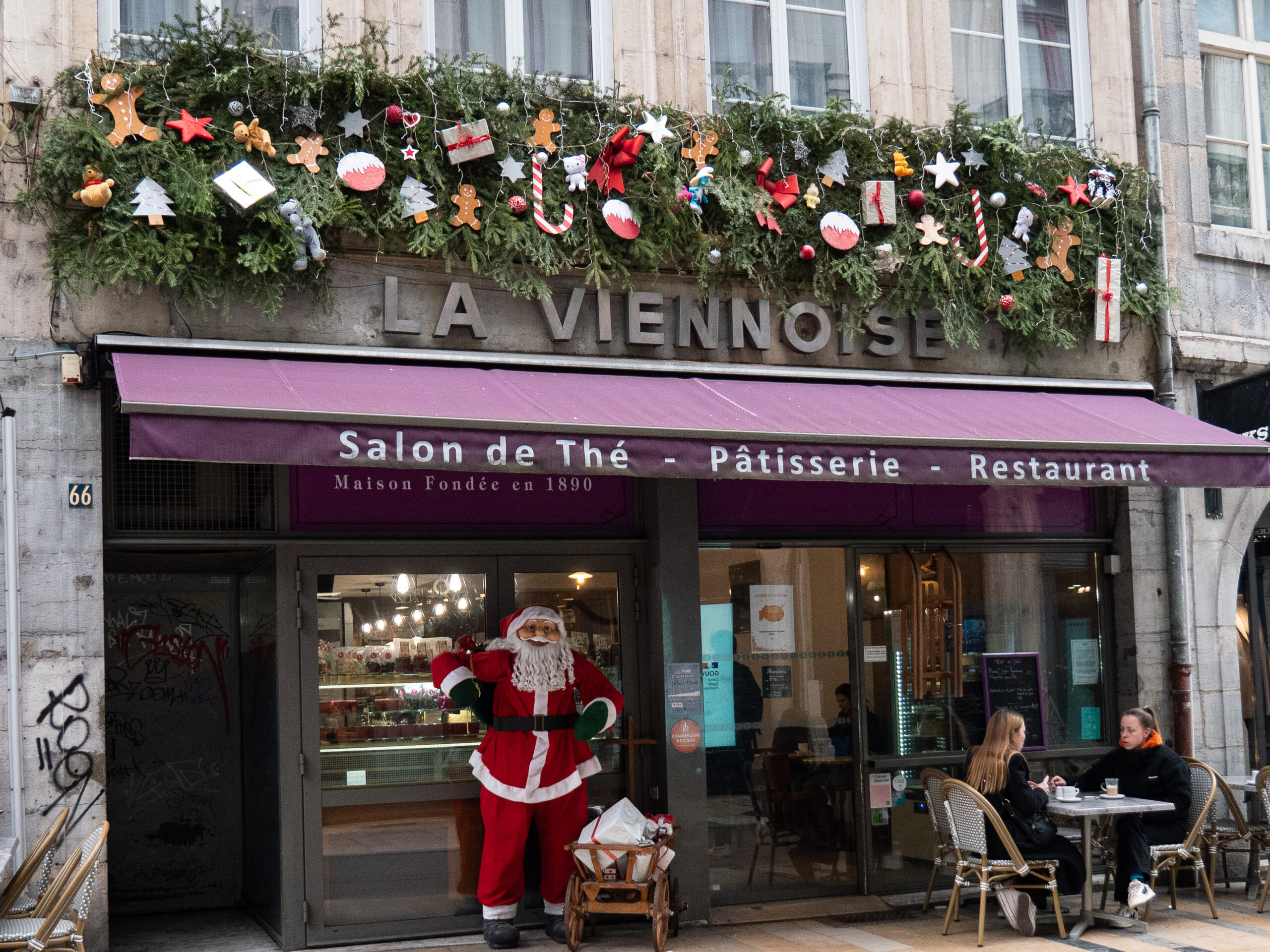 Une guirlande faite de branches de sapin, divers cadeaux, bonbons et viennoiseries en bois, installée au-dessus d'une pâtisserie dans la grande rue de Besançon