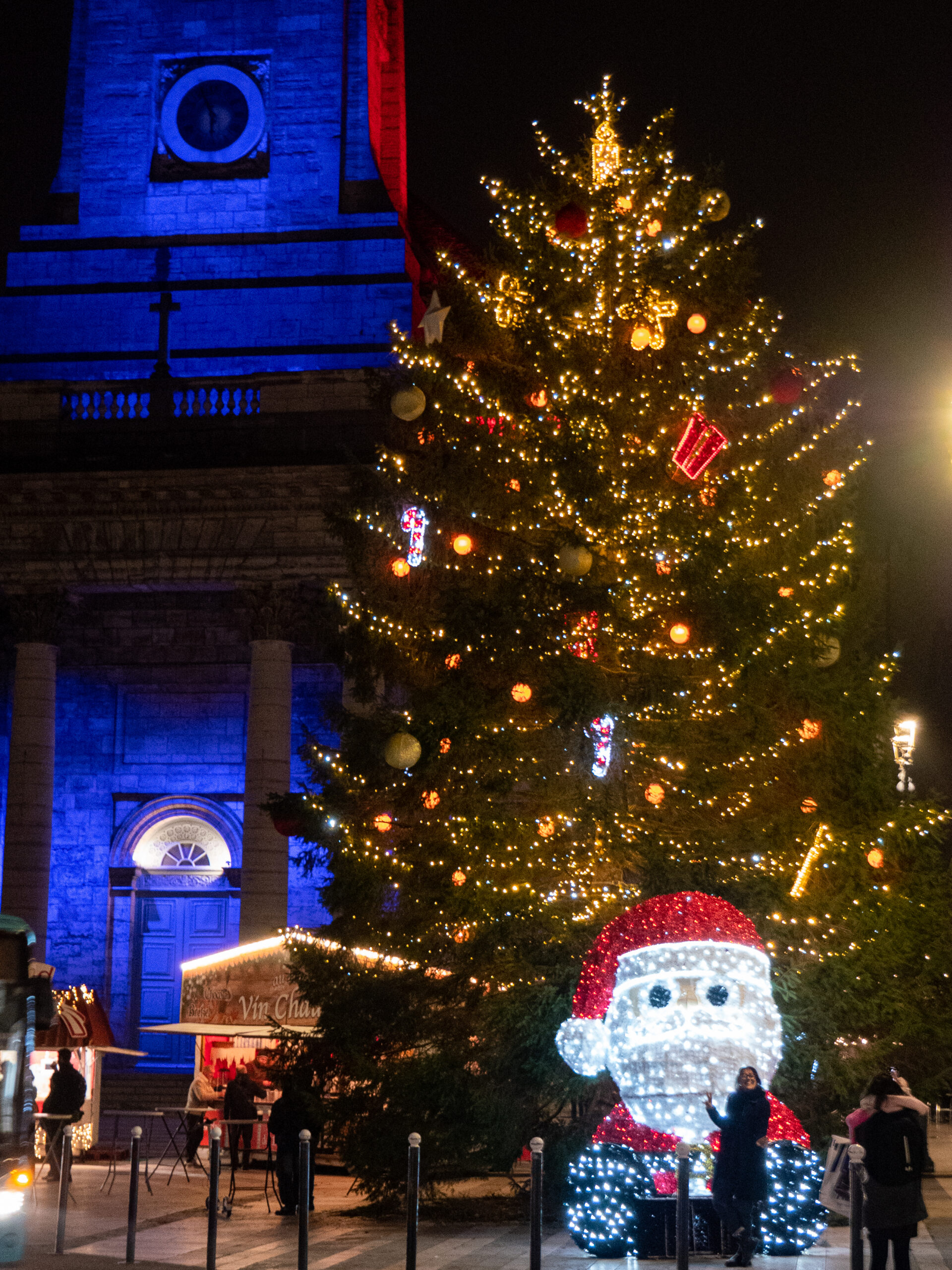 le grand sapin tout décoré de la place du 8 septembre, devant l'église éclairée de bleue. Il y a un père noël tout allumé de 2 mètres 50 devant le sapin et un stand de vin chaud derrière