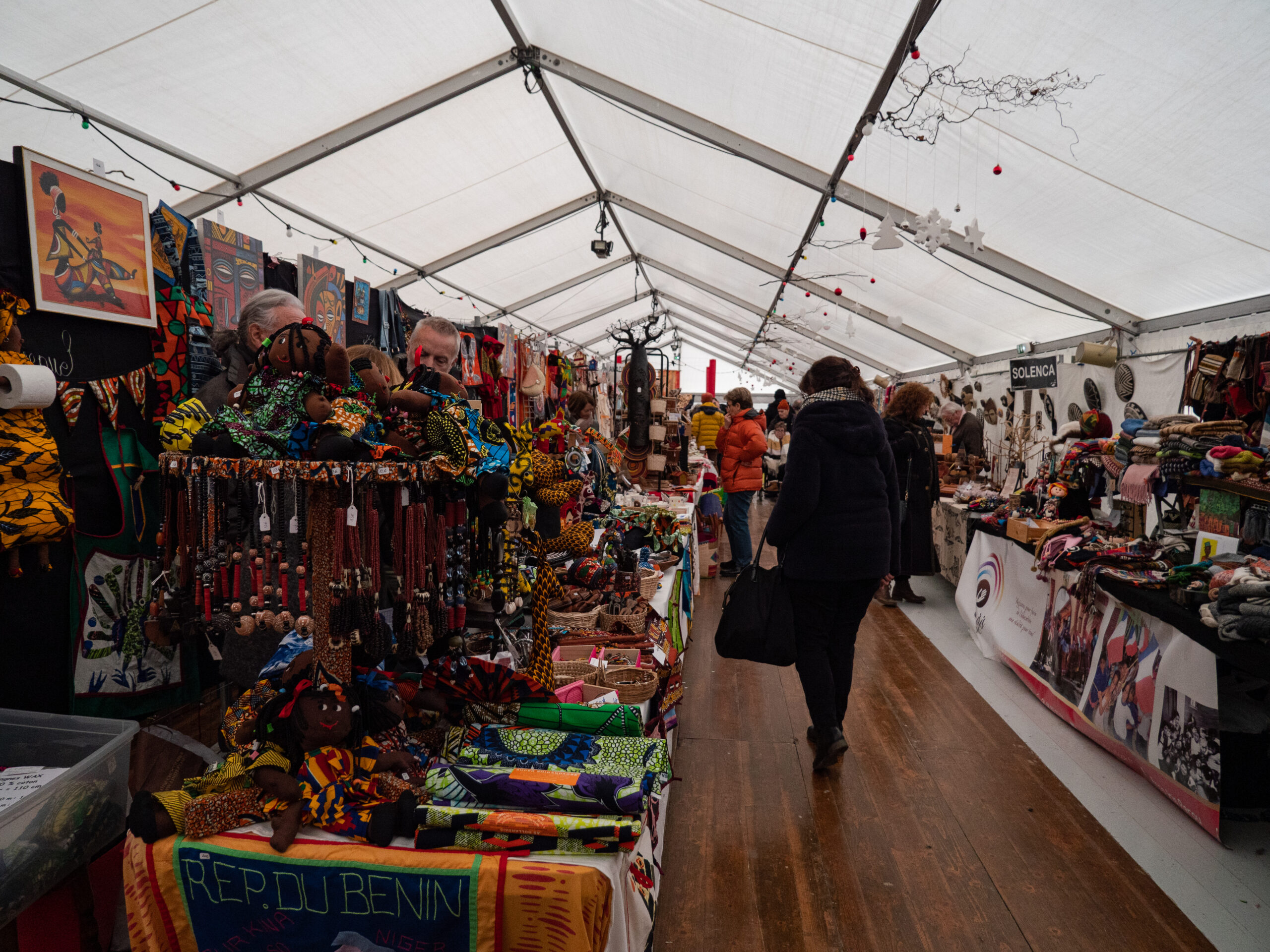 La même allée sous le long chapiteau du marché de Noël solidaire, pleine de stands de tissus, objets artisanaux en bois ou en cuir, devant lesquels quelques personnes sont arrêtées. On y voit plus spécifiquement les fabrications du bénin : poupées artisanales, tissus, bijoux et broches en bois