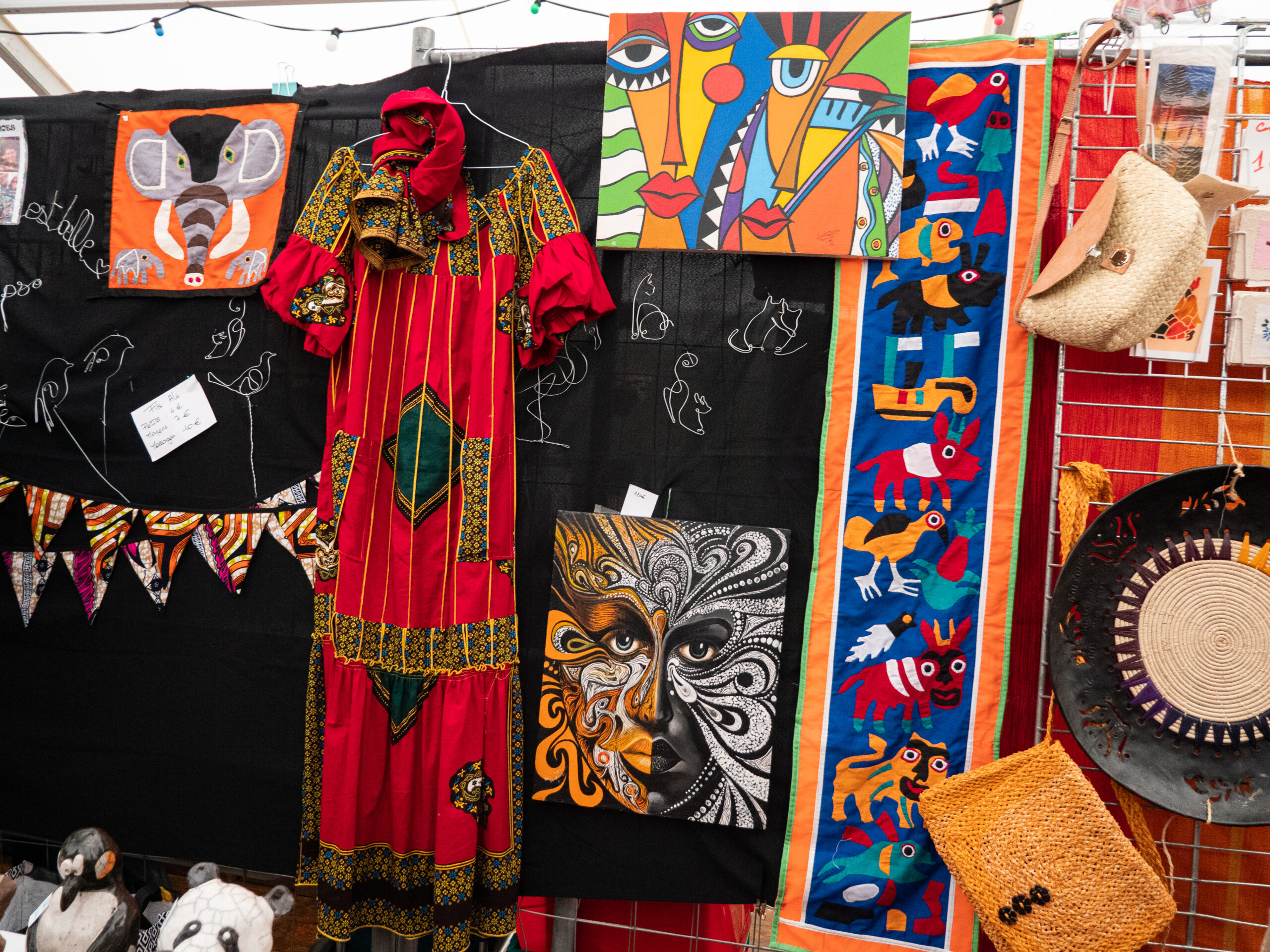 Stand avec de multiples produits : boubou très coloré à dominante rouge, tableau noir, blanc et jaune représentant un visage, étoffe avec des animaux stylisés, sculptures animales en fil d'alu, sacs à main tressé, fanions