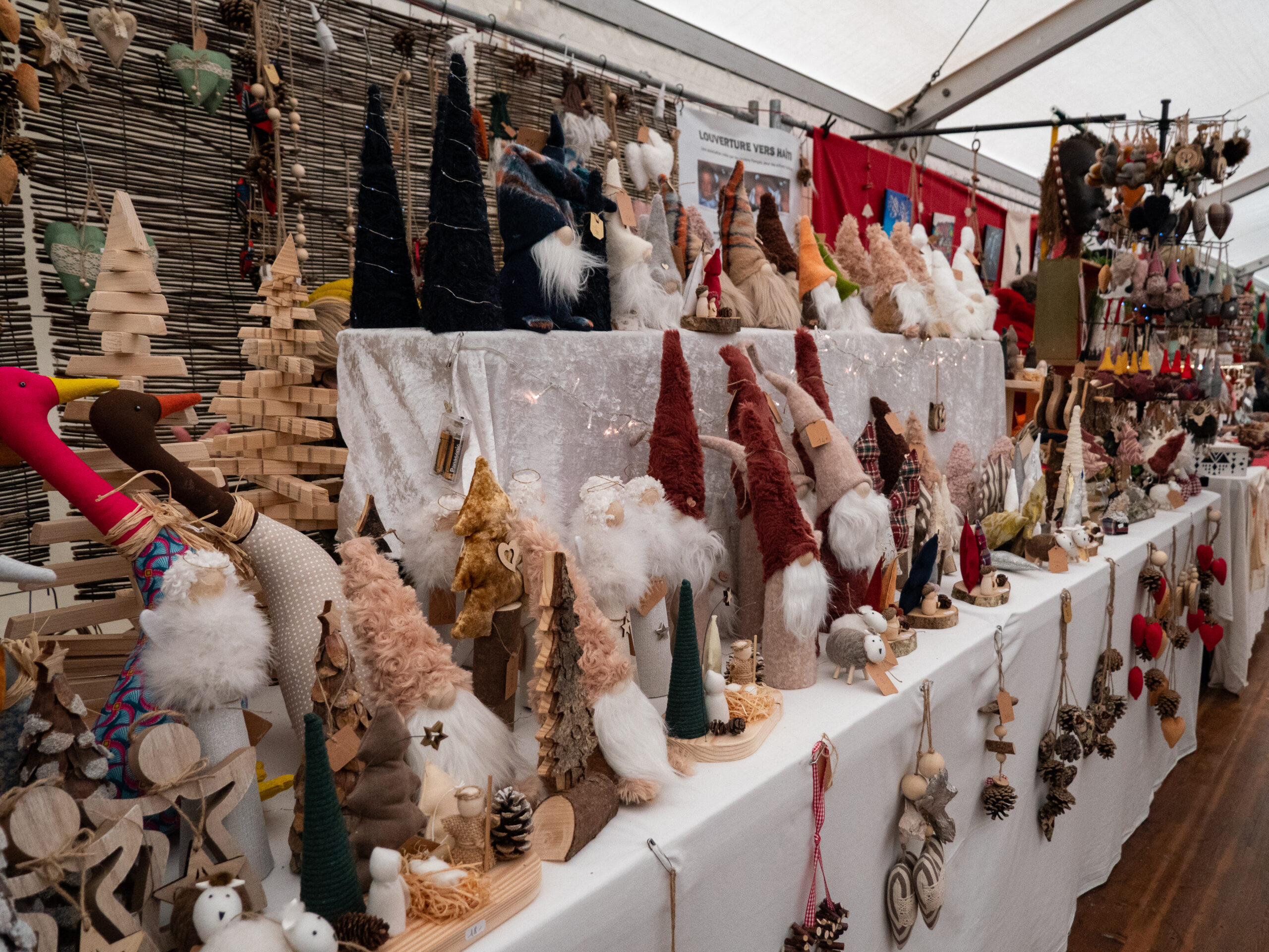 Stand garni de créations en tissus, telles que des lutins à bonnet haut, des moutons tout mignons, des pendentifs en bois et pommes de pin, représentant parfois des animaux, le tout présenté sur un grand linge blanc le long de l'allée