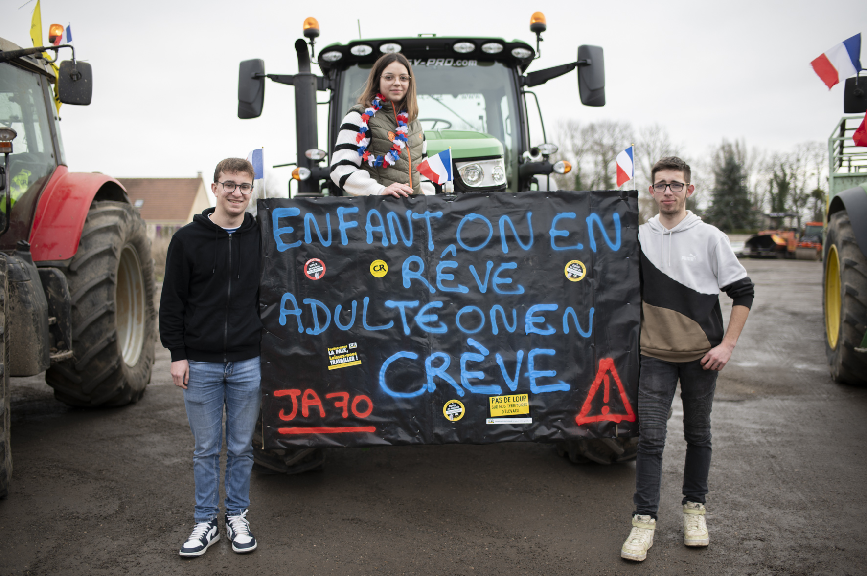 Une banderole dépliée devant un tracteur, avec deux jeunes hommes sur les flancs et une jeune femme en haut. On y lit : enfant on en rêve, adulte on en crève, signé "Jeunes Agriculteurs 70"