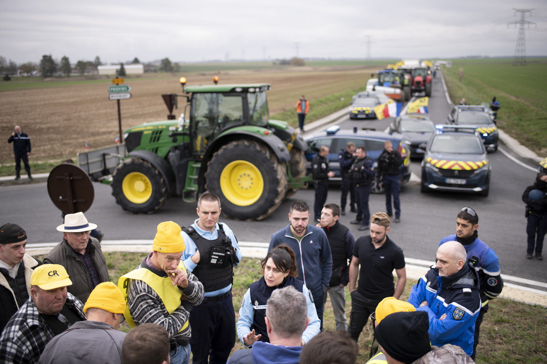 Réunion de plusieurs personnes sur un rond point, encadrée par la police. La route est bloquée par les tracteurs et les voitures de police.