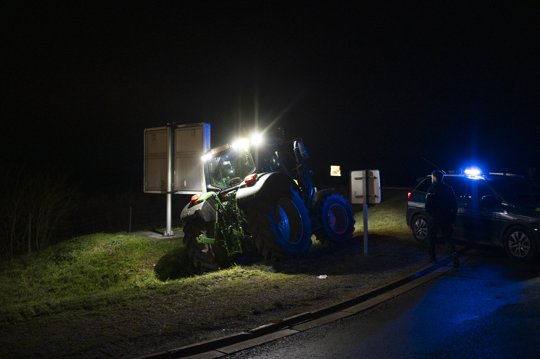 Photo de nuit, un tracteur tout éclairé à côté d'un panneau routier, sur le bas côté, à côté d'une voiture de police