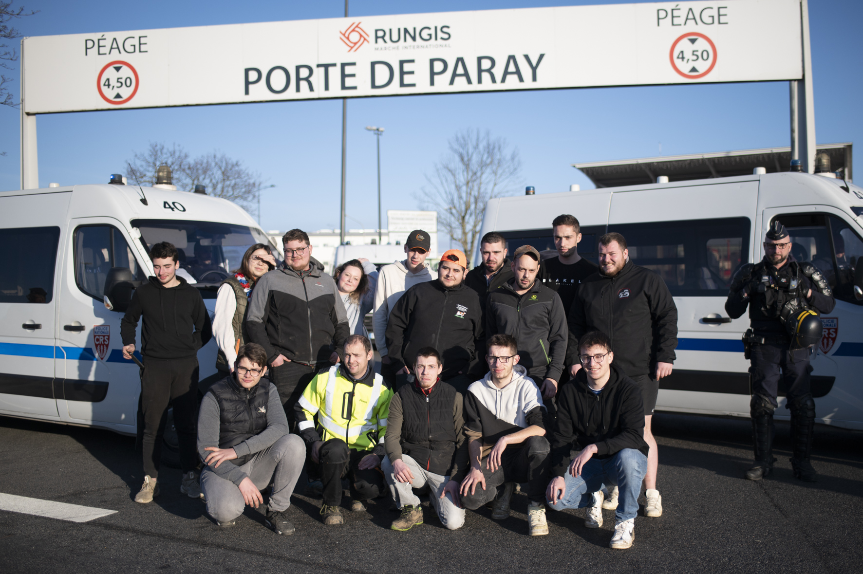 une douzaine de jeunes agriculteurices en train de prendre la pose, une rangée debout une assise, sous la porte de Paray de Rungis, encadrés par la police