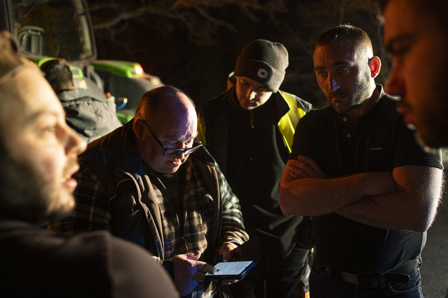 Photo de nuit, 3 hommes consultent un smartphone et discutent, avec deux têtes d'homme en avant plan et un tracteur en fond