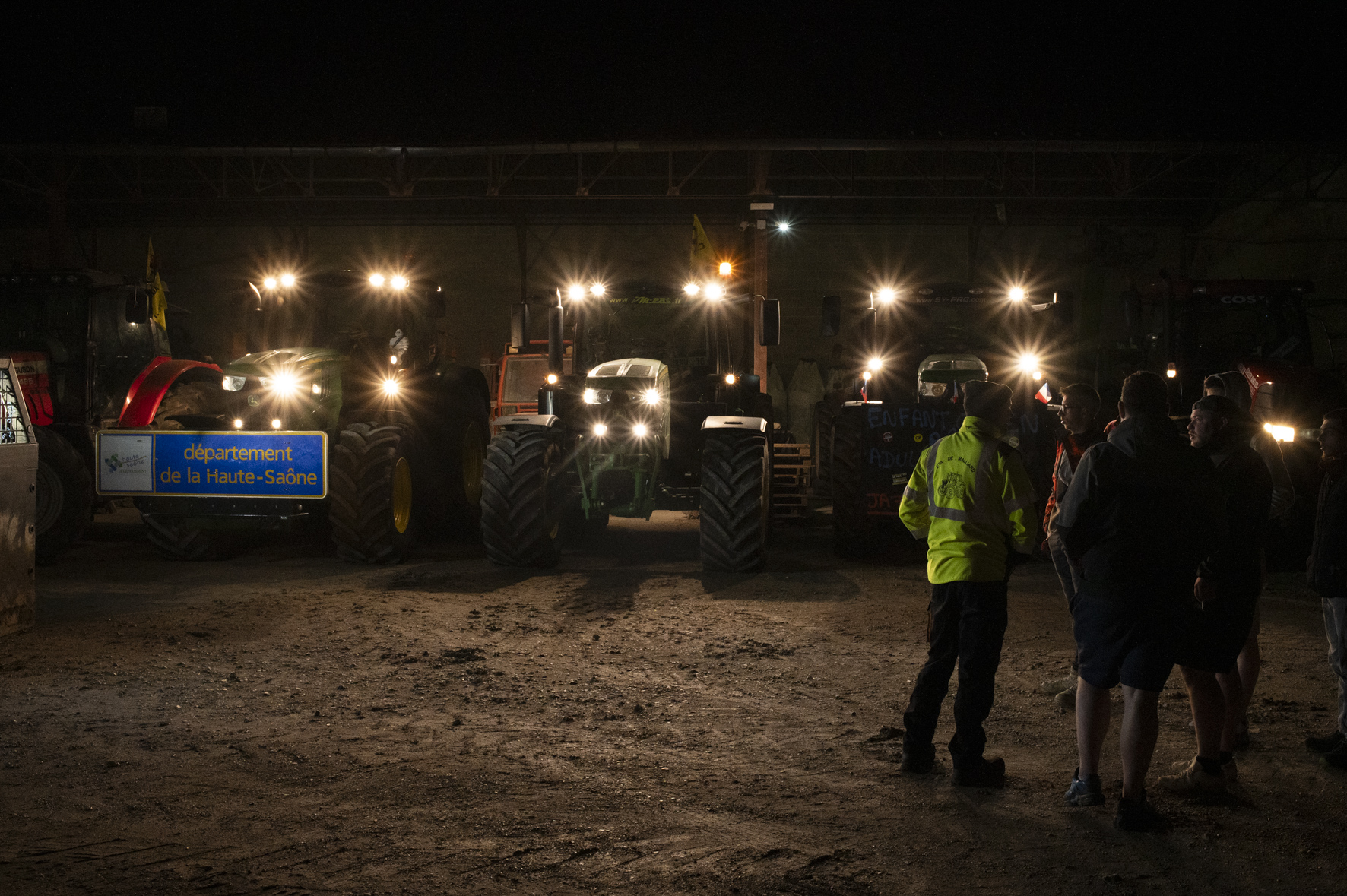 photo de nuit, cinq homme sont réunis à droit de l'image, devant une rangée de tracteurs garés sous une structure métallique, tous phares allumés.