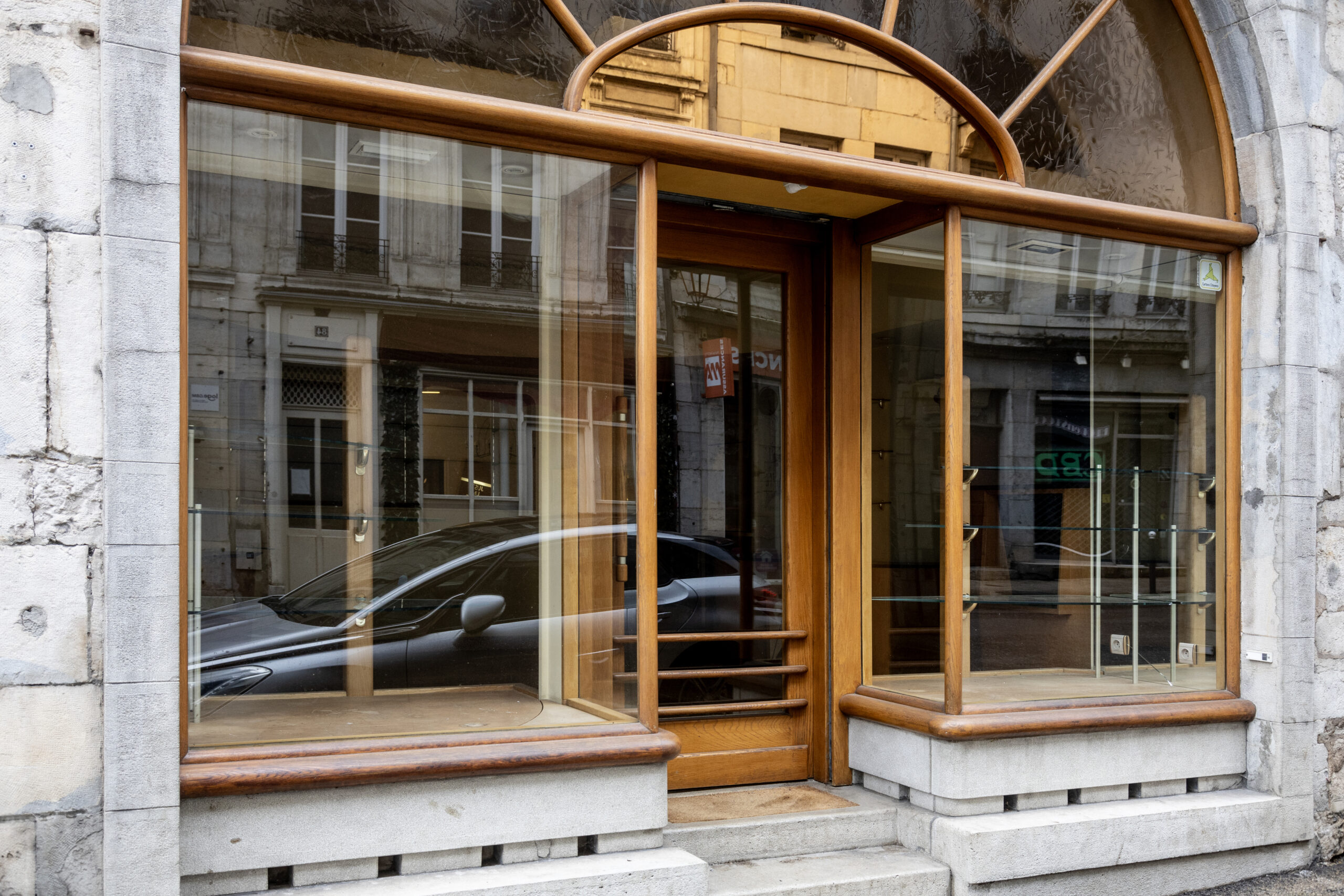 Photo de la devanture du chausseur Mouillebec, magasin centenaire, fermé, les vitrines et la porte aux montants boisés laissées visibles, intérieur vide.