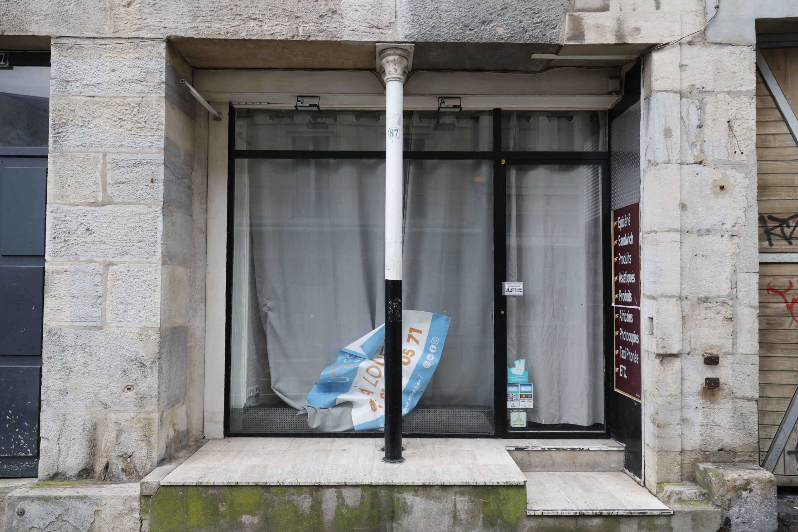 Photo de la devanture d'épicerie, fermée, avec un rideau pour opacifier l'intérieur et un panneau de location tombé derrière la vitrine