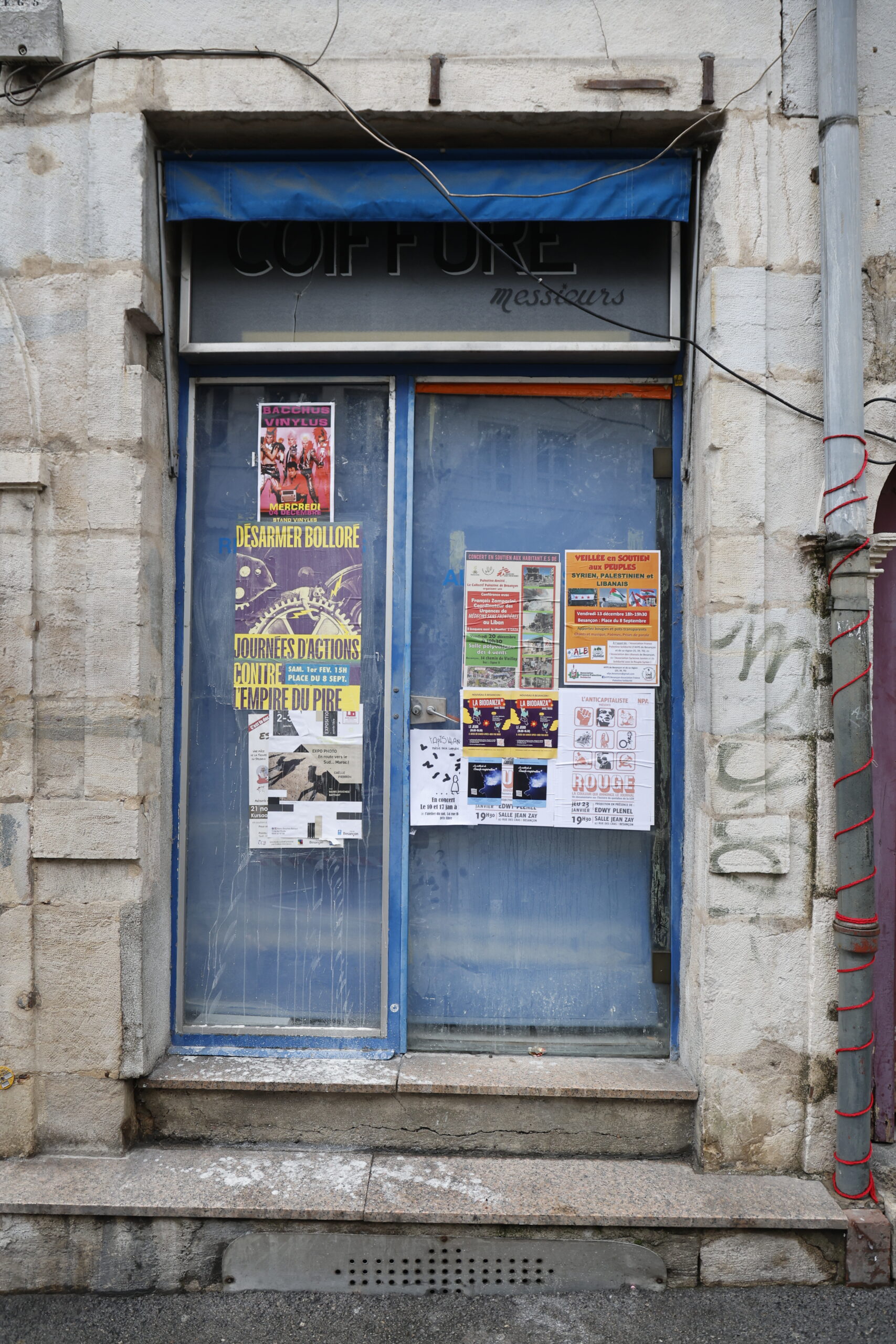 Photo de la devanture d'un salon de coiffure, fermé, sa petite vitrine au rideau bleu entièrement tiré recouverte d'affiches