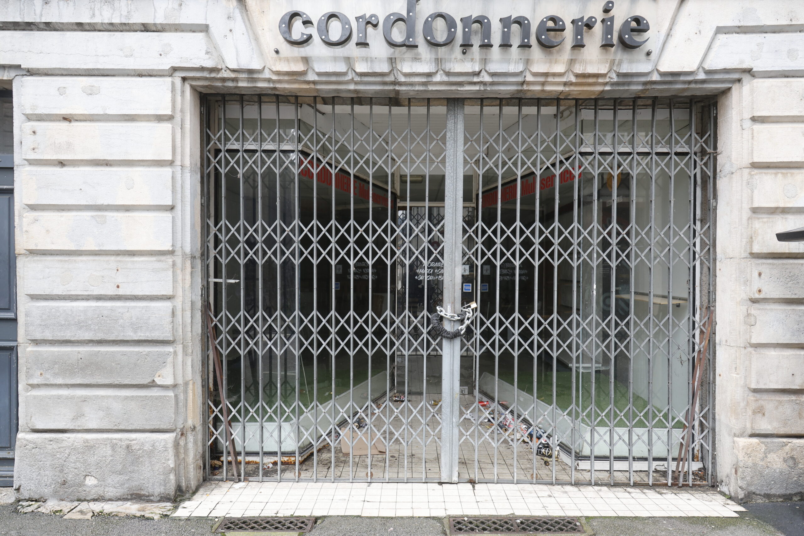 Photo de la devanture d'une cordonnerie, fermée, les locaux laissés vides, de nombreux déchets ayant été jetés entre le rideau de fer et la vitrine