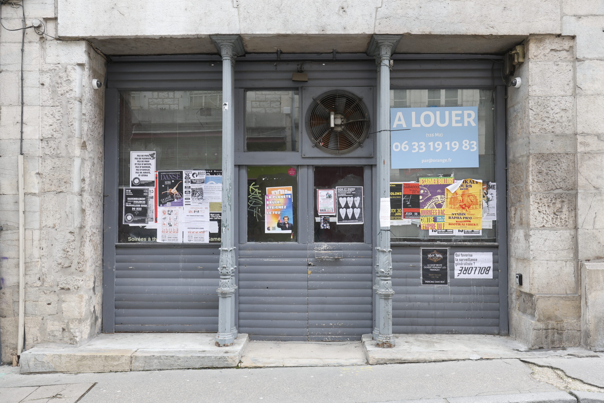 Photo de la devanture de magasin inconnu, fermé, les portes grises fermées et recouvertes de nombreuses affiches, ainsi que d'un panneau "à louer"