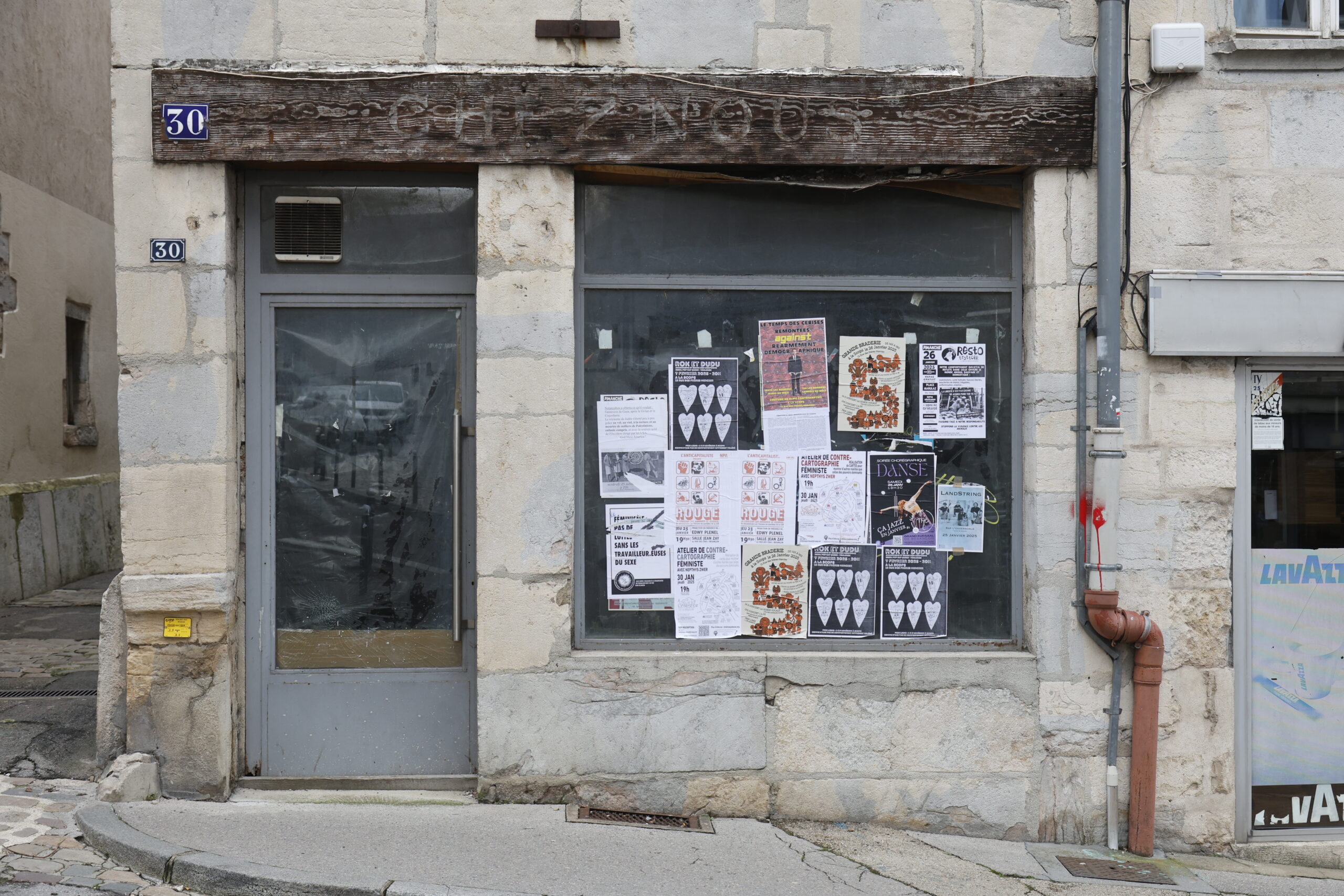 Photo de la devanture d'un commerce inconnu, fermé. La poutre au-dessus de l'entrée indique encore "CHEZ NOUS" en lettres effacées, la vitrine et la portée opacifiées au noir, avec de nombreuses affiches sur la vitrine