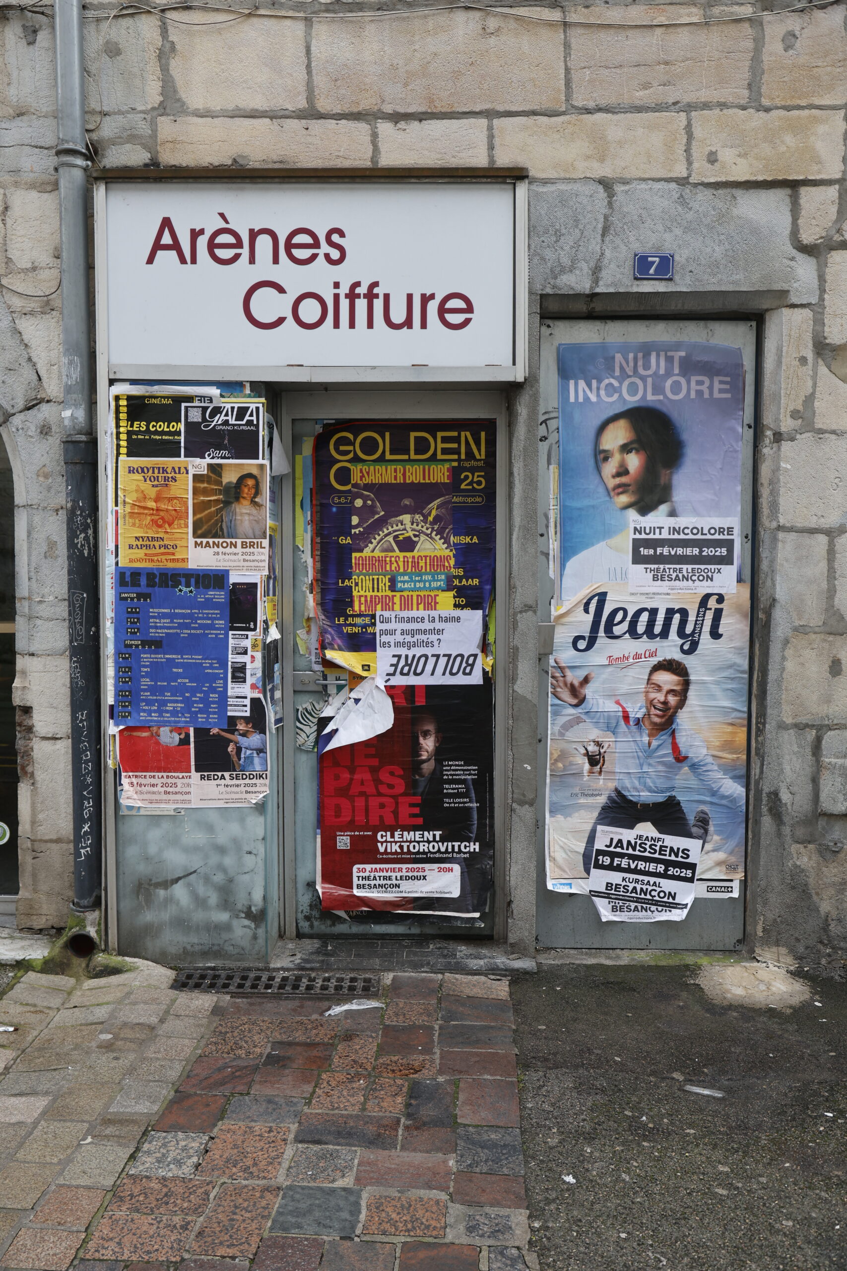 Photo de la devanture du salon Arènes Coiffure, fermé, porte et vitrine recouvertes de nombreuses affiches