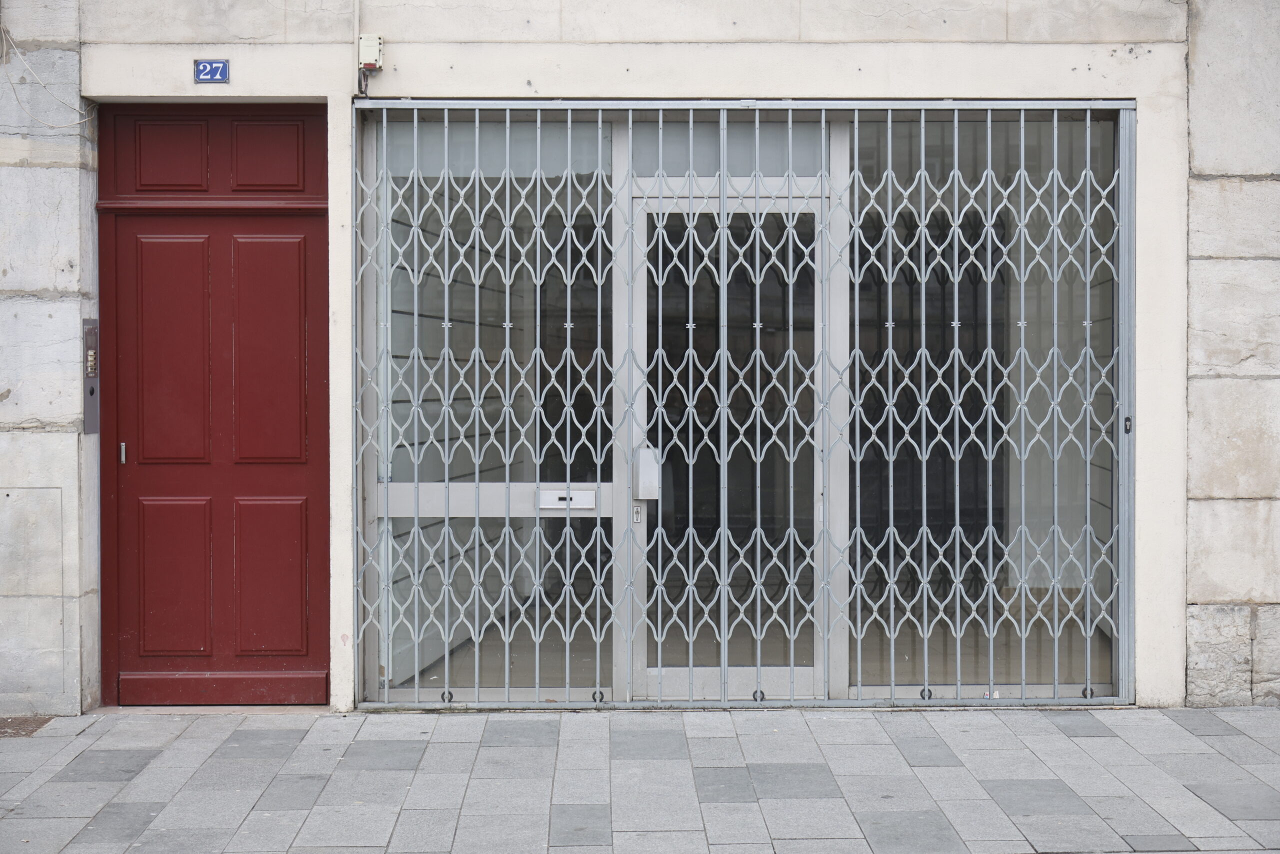 Photo d'une devanture inconnue, le grillage fermant entièrement la vitrine et la portée vitrée, à côté d'une porte rouge.