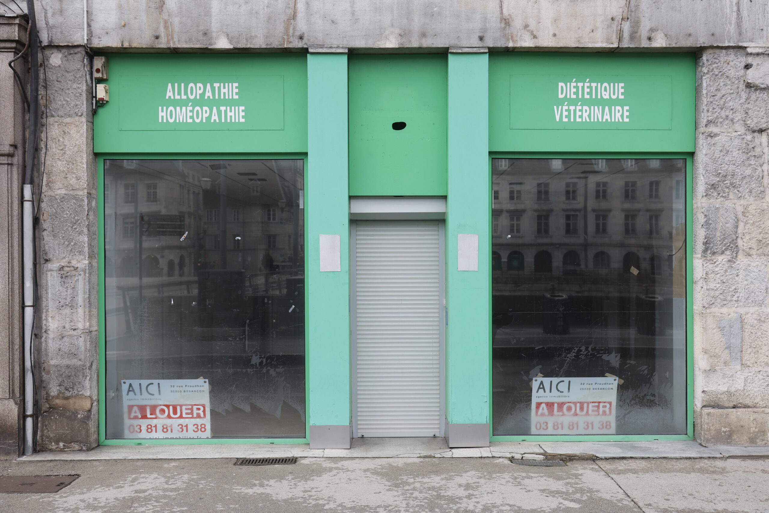 Photo de la devanture d'une pharmacie, fermée, le rideau de fer baissé et la vitrine entièrement vidée, panneaux "à louer" bien en évidence