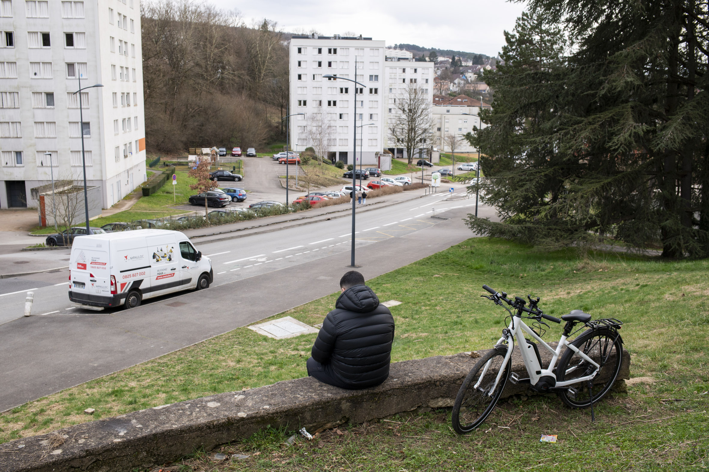 Un jeune homme de dos assis sur un muret, son vélo est a côté de lui, il est en hauteur par rapport à la rue de Chalezeule